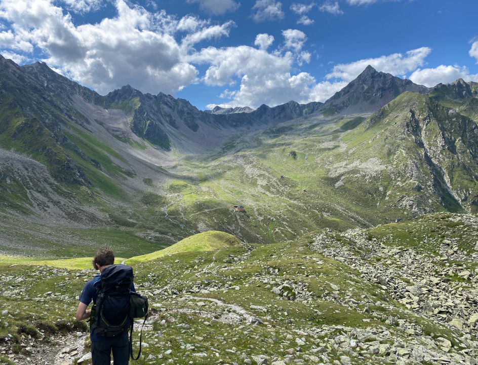 Uitzicht op groene bergweides met wat stenen, waarbij je over de schouder van een wandelaar kijkt